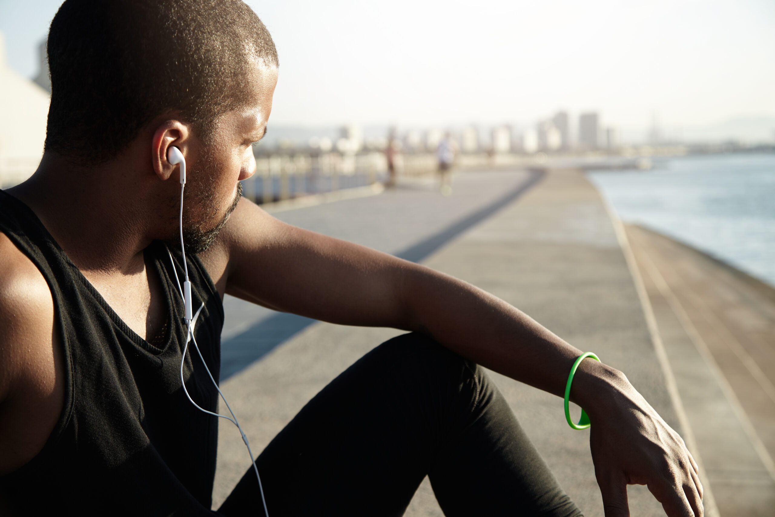Cropped portrait of young African man with muscular body wearing earphones while resting alone on embankment. Handsome athlete in black tank top listening to music with headphones relaxing outdoors